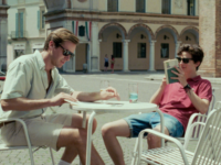 Cena do filme Me Chame pelo seu Nome que mostra Elio e Oliver sentados em uma mesa de café em uma praça.