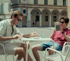 Cena do filme Me Chame pelo seu Nome que mostra Elio e Oliver sentados em uma mesa de café em uma praça.