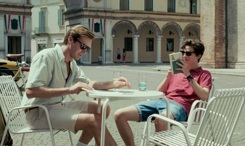 Cena do filme Me Chame pelo seu Nome que mostra Elio e Oliver sentados em uma mesa de café em uma praça.