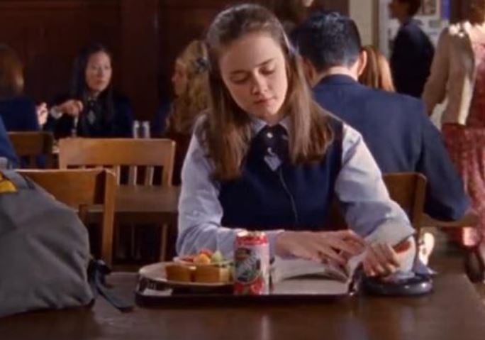 A personagem Rory almoçando sozinha na escola enquanto lê um livro, na série Gilmore Girls. 